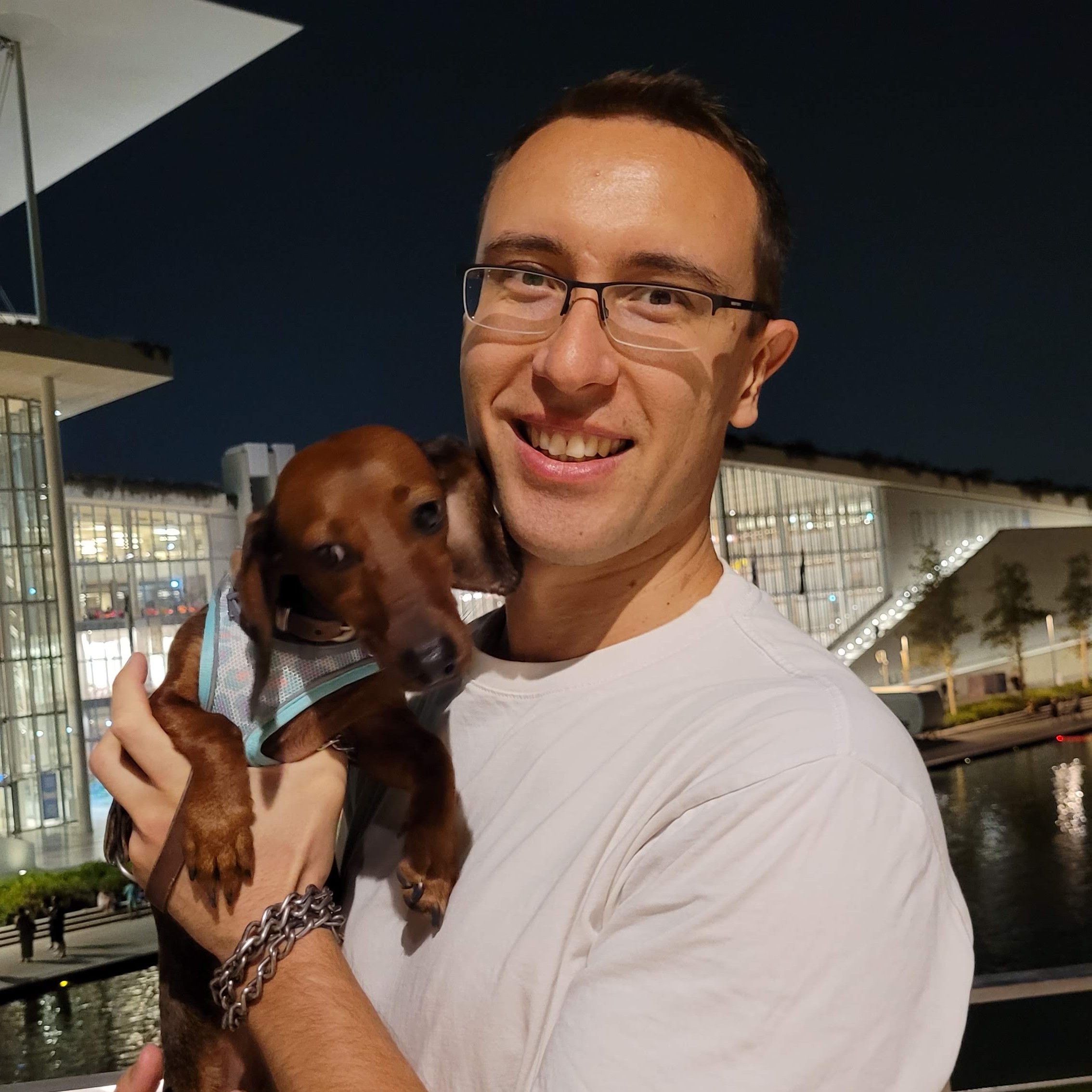 Alexander Kyriakou holding a dachshund dog in front of Stavros Niarchos Foundation Cultural Center in the summer of 2024